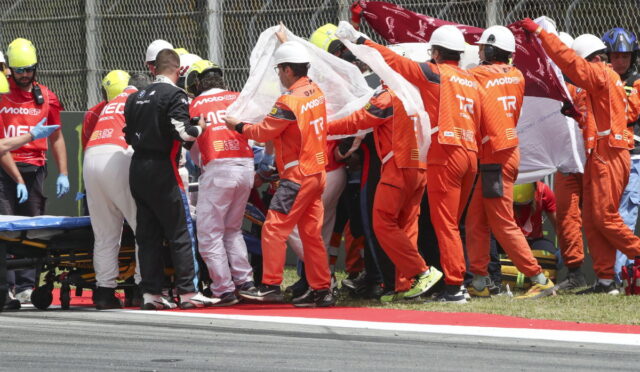 Pit crew in orange suits and medical staff carry a white sheet over a stretcher beside a crash on the track.