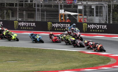 Group of motorcycle racers leaning into a sharp turn on a race track, Monster Energy banners in the background.