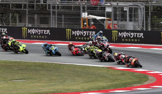 Group of motorcycle racers leaning into a sharp turn on a race track, Monster Energy banners in the background.