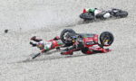 Two motorcycle racers crash on a gravel track; red-and-white bikes tumble as riders slide across the gravel.