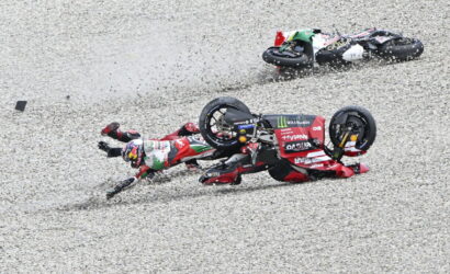 Two motorcycle racers crash on a gravel track; red-and-white bikes tumble as riders slide across the gravel.