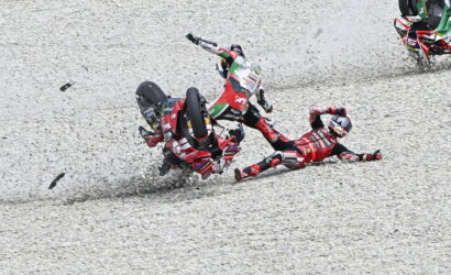 Motocross crash on a gravel track: two riders in red gear fall while a third rider clips by, kicking up rocks.
