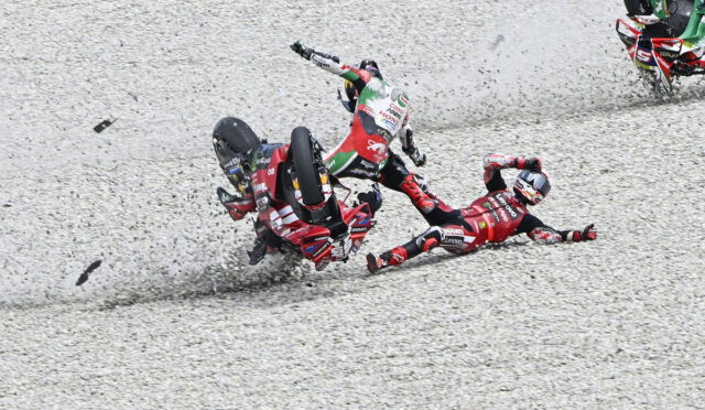 Motocross crash on a gravel track: two riders in red gear fall while a third rider clips by, kicking up rocks.