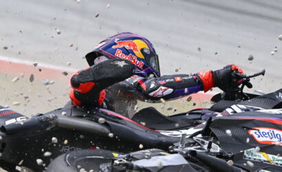 Racing motorcyclist in a Red Bull helmet slides into a pile-up, gravel and bike parts flying around.