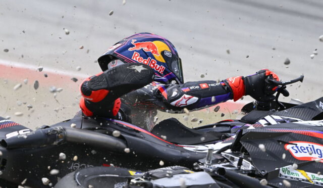 Racing motorcyclist in a Red Bull helmet slides into a pile-up, gravel and bike parts flying around.