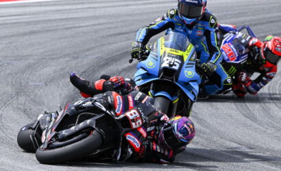 Three motorcycle racers lean into a sharp turn on a racing track; blue bike leads, black bike close behind, all in full racing gear on slick tires.