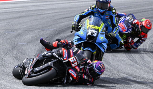 Three motorcycle racers lean into a sharp turn on a racing track; blue bike leads, black bike close behind, all in full racing gear on slick tires.