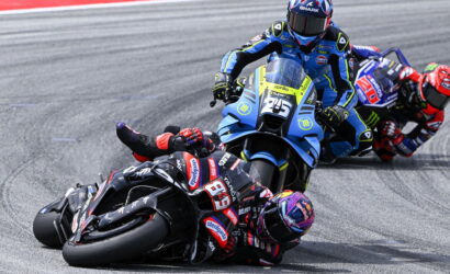 MotoGP riders lean into a left-hand corner, foreground rider on a black bike #93 with red accents, blue-yellow bike close behind.
