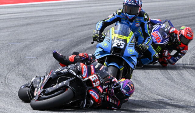 MotoGP riders lean into a left-hand corner, foreground rider on a black bike #93 with red accents, blue-yellow bike close behind.