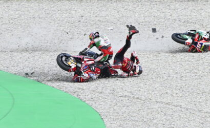 Two motorcycle racers crash on a gravel corner; one rider lies on the ground with a leg raised as bikes skid nearby.