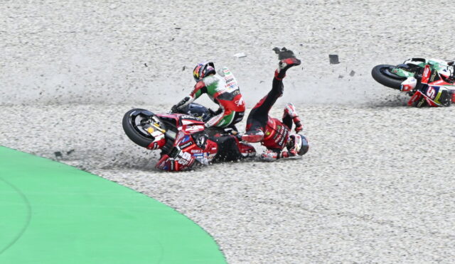 Motorcycle crash on a race track: two riders in red gear fall onto gravel with bikes skidding and debris around them.