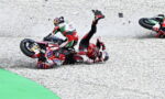 Two motorcycle racers crash on a gravel run-off, bikes skidding and debris in the air as riders tumble.
