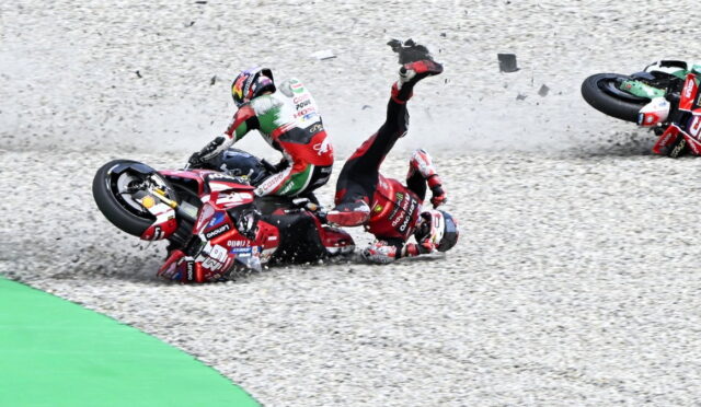 Two motorcycle racers crash on a gravel run-off, bikes skidding and debris in the air as riders tumble.