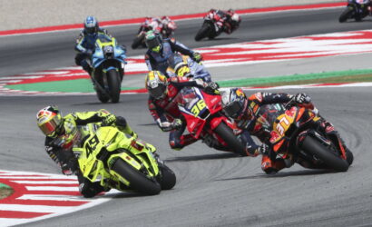 Group of motorcycle racers lean into a sharp turn on a race track, front rider in bright neon yellow suit and bike leading the pack.
