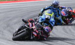 Motorcycle racers lean into a tight turn on a racetrack; lead rider #93 in red–black gear, blue and yellow bikes close behind.