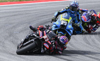 Motorcycle racers lean into a tight turn on a racetrack; lead rider #93 in red–black gear, blue and yellow bikes close behind.