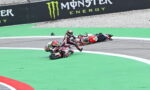 Two motorcycle riders crash on a race track: one bike upright, the other on its side near the green painted area, with a fallen rider nearby and a Monster Energy barrier in the background.