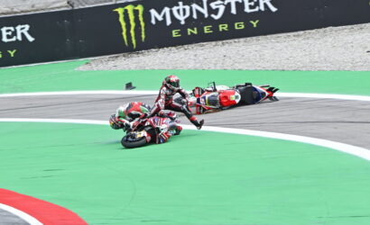 Two motorcycle riders crash on a race track: one bike upright, the other on its side near the green painted area, with a fallen rider nearby and a Monster Energy barrier in the background.