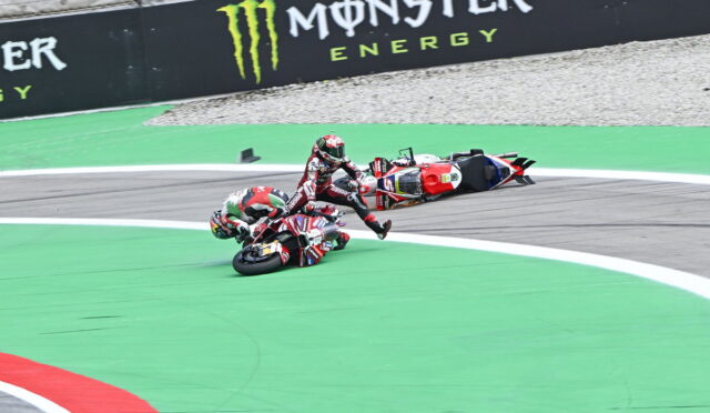 Two motorcycle riders crash on a race track: one bike upright, the other on its side near the green painted area, with a fallen rider nearby and a Monster Energy barrier in the background.