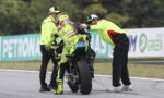 Racer in neon yellow gear being helped by crew as they stabilize the bike on the track edge.