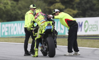 Racer in neon yellow gear being helped by crew as they stabilize the bike on the track edge.