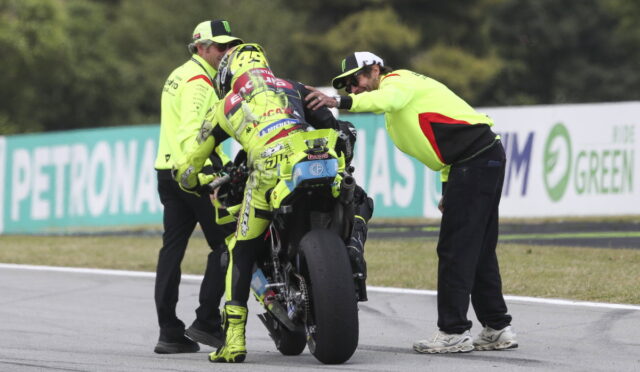 Racer in neon yellow gear being helped by crew as they stabilize the bike on the track edge.