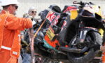Damaged racing motorcycle on a lift, being recovered as orange-clad marshals observe behind a chain-link fence.