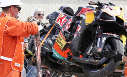 Damaged racing motorcycle on a lift, being recovered as orange-clad marshals observe behind a chain-link fence.