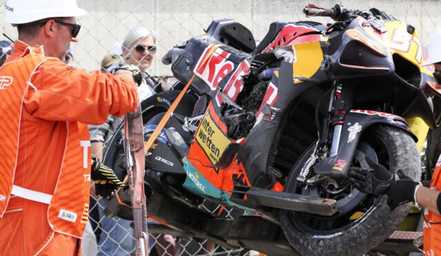 Damaged racing motorcycle on a lift, being recovered as orange-clad marshals observe behind a chain-link fence.