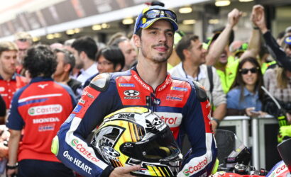 Motorcycle racer in blue-red suit holds a colorful helmet, standing in a crowded pit area with cheering fans behind him