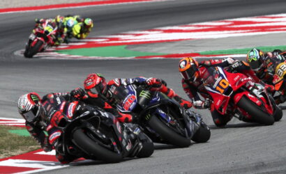 MotoGP riders lean into a tight corner, racing on a colorful track with red, white, and green curbs visible in the background.