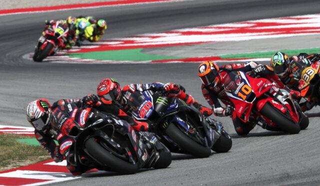 MotoGP riders lean into a tight corner, racing on a colorful track with red, white, and green curbs visible in the background.