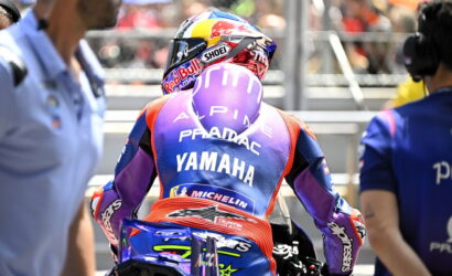 Racer from behind in a purple Yamaha Pramac racing suit sitting on a blue motorcycle in the pit lane, helmet on.