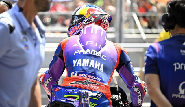 Racer from behind in a purple Yamaha Pramac racing suit sitting on a blue motorcycle in the pit lane, helmet on.
