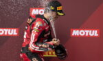 Racing driver in red suit sprays champagne on podium to celebrate a win, holding a helmet while water droplets fly around.