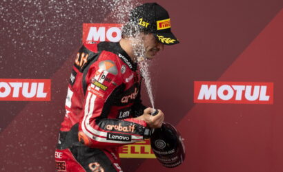 Racing driver in red suit sprays champagne on podium to celebrate a win, holding a helmet while water droplets fly around.