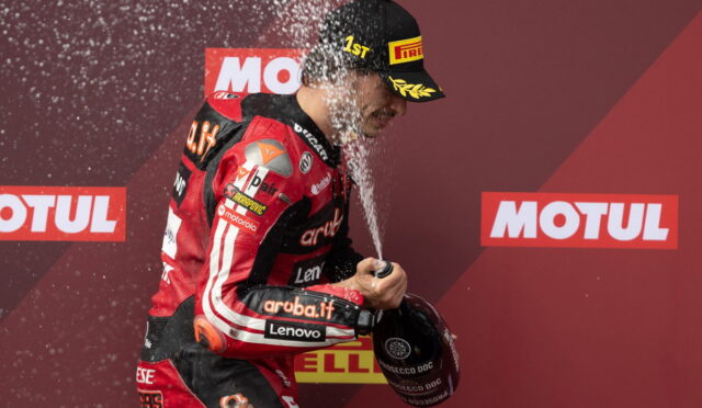 Racing driver in red suit sprays champagne on podium to celebrate a win, holding a helmet while water droplets fly around.