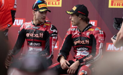 Two motorcycle racers in red Aruba.it racing suits sit and chat on a stage with Pirelli banners in the background.