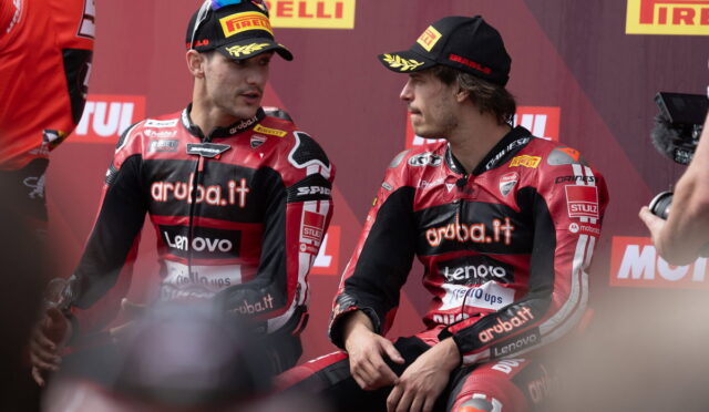 Two motorcycle racers in red Aruba.it racing suits sit and chat on a stage with Pirelli banners in the background.