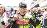 Moto racer in a neon Enduro suit, wearing a blue cap and sunglasses, holds a Monster Energy drink and points upward in a busy pit lane.