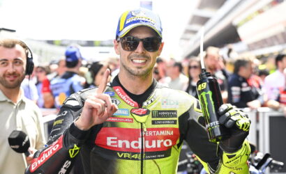 Moto racer in a neon Enduro suit, wearing a blue cap and sunglasses, holds a Monster Energy drink and points upward in a busy pit lane.