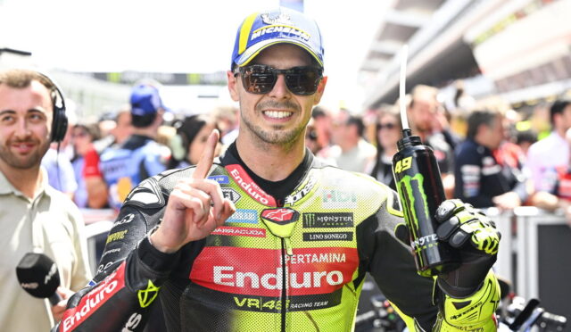 Moto racer in a neon Enduro suit, wearing a blue cap and sunglasses, holds a Monster Energy drink and points upward in a busy pit lane.