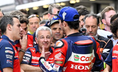 MotoGP rider in red/blue Honda gear addresses a crowd of fans and reporters (back view). Best viewed from behind with Castrol Honda branding visible.