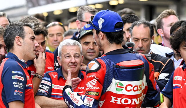 MotoGP rider in red/blue Honda gear addresses a crowd of fans and reporters (back view). Best viewed from behind with Castrol Honda branding visible.