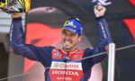 Motorsport racer on a podium celebrating victory with arms raised, wearing a blue-red Honda racing suit and a Michelin cap, holding a helmet overhead.