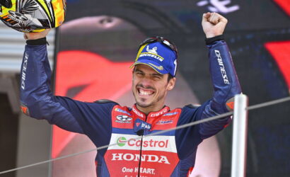 Motorsport racer on a podium celebrating victory with arms raised, wearing a blue-red Honda racing suit and a Michelin cap, holding a helmet overhead.