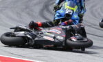 Racer in black suit and red gloves lies beside a black motorcycle on a curving race track; a blue bike closes in from behind.