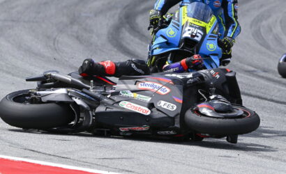 Racer in black suit and red gloves lies beside a black motorcycle on a curving race track; a blue bike closes in from behind.