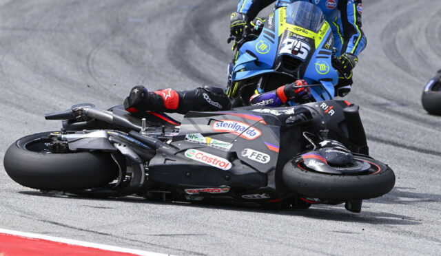Racer in black suit and red gloves lies beside a black motorcycle on a curving race track; a blue bike closes in from behind.