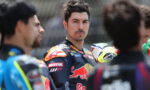 Close-up of a racing rider with dark hair and stubble, wearing a Red Bull KTM racing suit, among teammates.